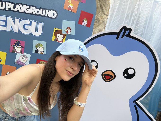 A young woman wearing a light blue cap and a heart on her cheek poses next to a large penguin mascot at what appears to be a 'Pengsoo Playground' event, with a backdrop featuring various character illustrations.