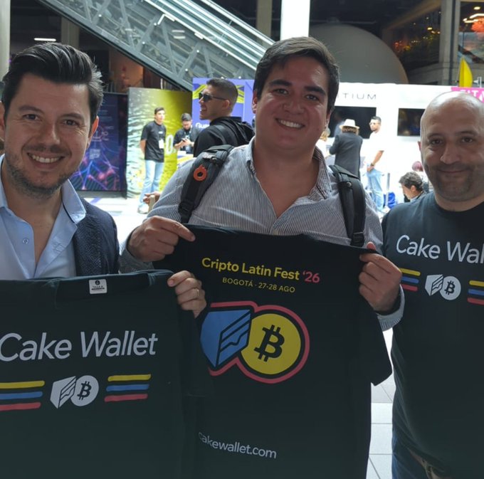 Three men smiling at a crypto event, holding branded merchandise for 'Cake Wallet' and 'Cripto Latin Fest '26' in Bogotá (27-28 August).