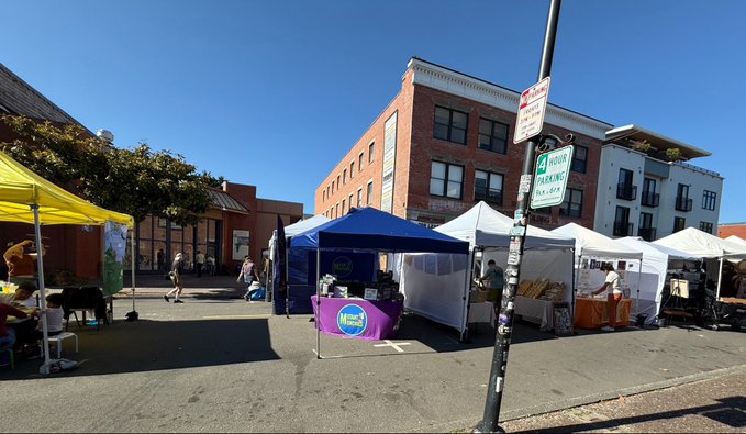 An outdoor street market scene with various vendor tents, including yellow, blue, and white canopies, set up along a street with brick buildings.