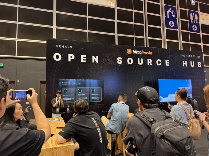 A photo taken at a Bitcoin Asia event showing an 'OPEN SOURCE HUB' booth with the numbers '~964479' and '103.144.202.255' displayed, where attendees are gathered around screens and wooden seating.