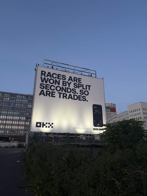 A large illuminated billboard displays the text 'RACES ARE WON BY SPLIT SECONDS. SO ARE TRADES.' alongside the OKX logo and an image of a smartphone, set against a twilight sky.