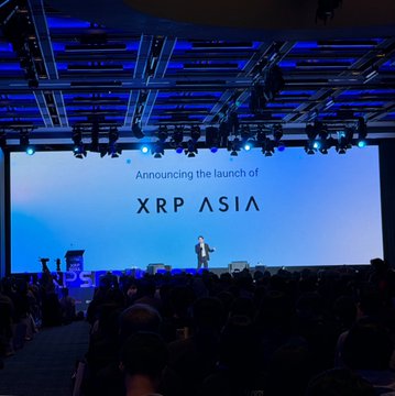 A presenter stands on a brightly lit stage in front of a large screen that reads 'Announcing the launch of XRP ASIA', addressing an audience.