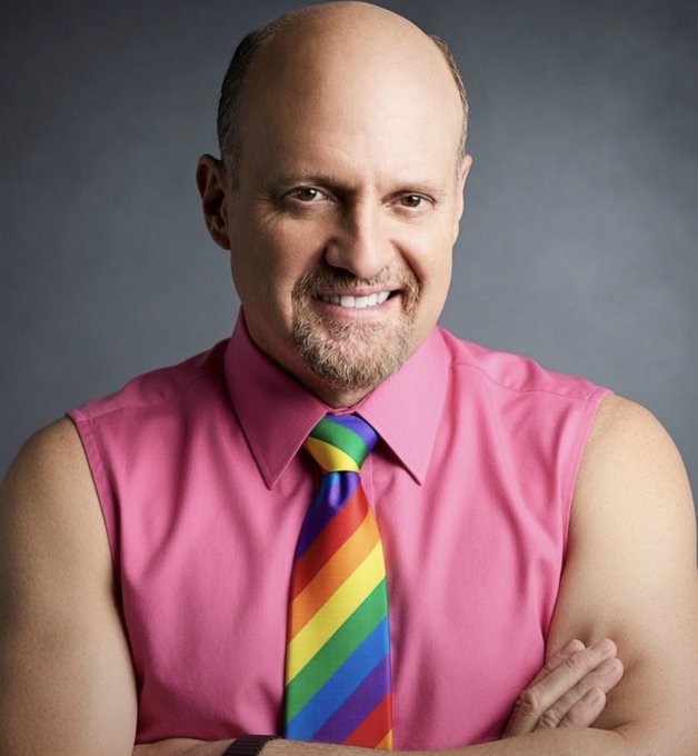 A professional portrait photograph of a smiling bald man with a goatee, wearing a pink sleeveless shirt and a colorful rainbow-striped tie, standing with arms crossed against a gray background.