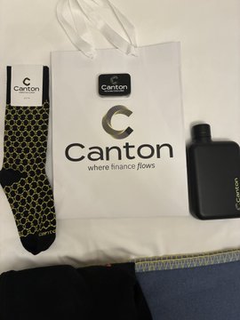 A gift bag and accessories from Canton, including socks, a black flask, and a tote bag with the "Canton" logo and tagline "where finance flows," likely received as a thank-you or swag bag from the Canton Summit.