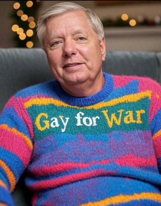 A man with gray hair sitting on a couch wearing a colorful striped sweater with the text 'Gay for War' knitted on it, with Christmas lights visible in the background.