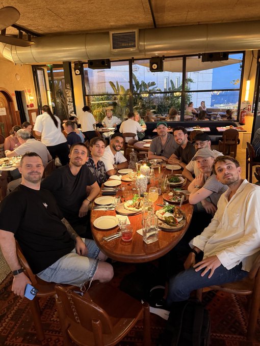 A group of about ten people sit around a long wooden table set with plates, glasses, and food at a lively restaurant, posing for the camera during a Turtle Group & friends dinner in Lisbon. The relaxed, smiling gathering captures the team catching up and connecting over a meal, matching the post's spirit of planning ahead and sharing exciting news.