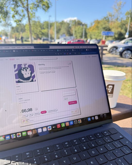 A laptop on a wooden table displays a profile page for 'zuzu' with an NFT of a chained character, showing a price of £66.98, with a Starbucks coffee cup and a parking lot in the background.