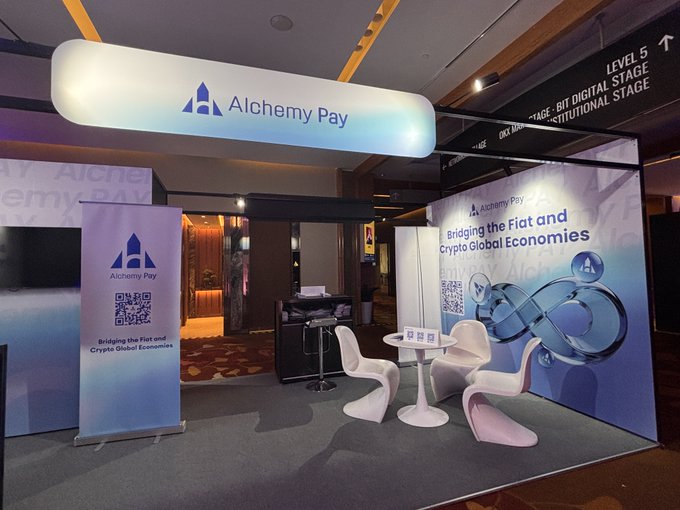A booth for 'Alchemy Pay' features illuminated signage with the tagline 'Bridging the Fiat and Crypto Global Economies', along with branded furniture and a QR code display.