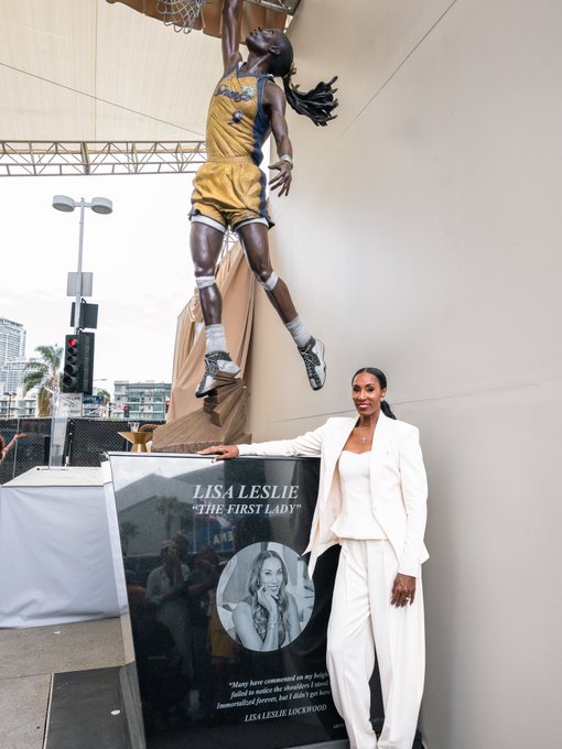 Lisa Leslie poses beside her newly unveiled statue at Crypto.com Arena's Star Plaza, with the display plaque reading "LISA LESLIE 'THE FIRST LADY'" and featuring the quote, "Many have commented on my height failed to notice that I dribbled, passed, Immortalized forever, but I didn't get here."