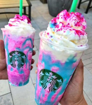 The image features two cups of a pink, blue, and white drink with rainbow-colored whipped cream toppings. One cup has a green straw sticking out of it, while the other has a yellow straw protruding from its top. The cups are placed on a table or sidewalk, suggesting that they might be enjoyed outdoors in an urban setting.