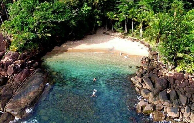 An aerial view of a secluded tropical beach cove with crystal-clear turquoise water, surrounded by lush green palm trees and rocky outcroppings, with a few people swimming and walking on the white sand.