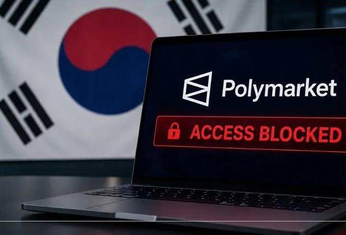 A laptop displays the Polymarket logo with a red "ACCESS BLOCKED" warning, set against a backdrop of the South Korean flag. The image illustrates South Korea's decision to block access to the Polymarket platform over concerns that it operates like an unlicensed gambling service.