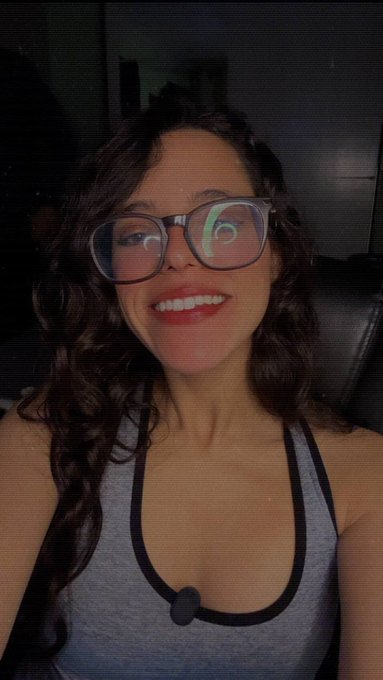 A slightly blurry selfie of a smiling young woman with long dark hair wearing glasses and a gray tank top, with a screen reflection visible in her lenses.