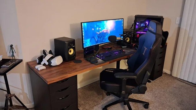 A home gaming/computer setup featuring a curved monitor displaying an underwater scene, a blue and black gaming chair, a VR headset, a keyboard with RGB lighting, and a desktop tower with purple lighting.