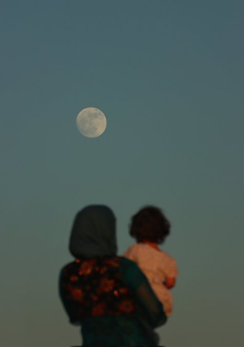 A silhouetted view from behind shows a person in a hooded floral garment holding a child, both gazing up at a full moon in a dusky blue sky.