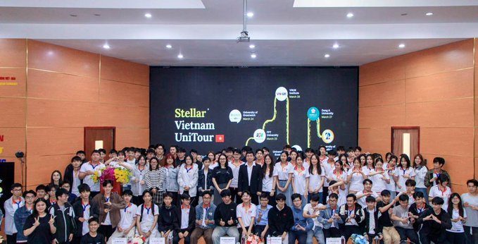 A large group of students and teachers pose together in an auditorium for a photo, with a projection screen behind them displaying the text 'Stellar Vietnam UniTour' alongside dates and event milestones.