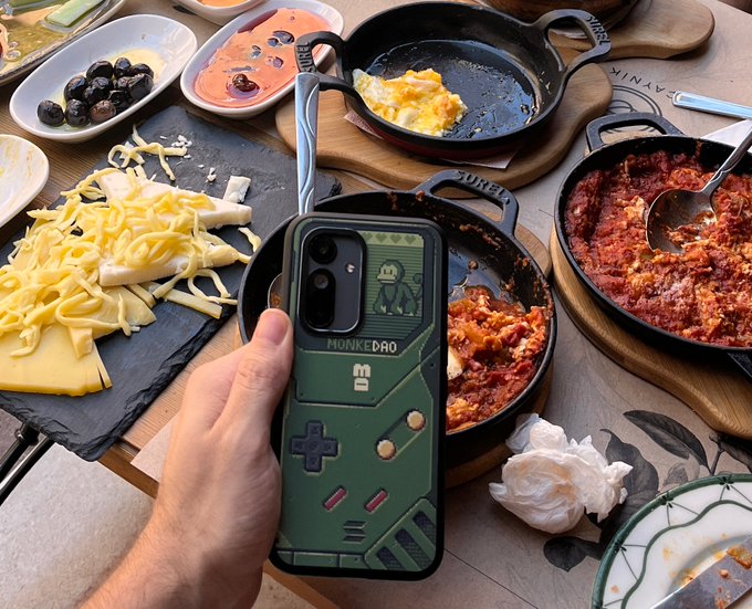 A hand holds a smartphone with a green phone case labeled "MONKE DAO" featuring a small monkey character, set against a background of assorted cheese, hot meals, and a dessert in cast-iron pans.
