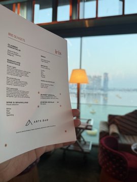 A hand holds up a printed 'IRIS' document labeled 'IRIS R3 SOKETS' (likely a menu or catalog) in a lounge area with a window view of a city skyline.