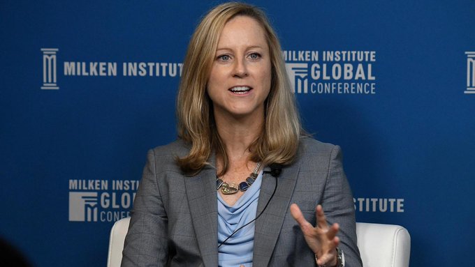 A blonde woman in a gray blazer speaks at the Milken Institute Global Conference, gesturing with her hand against a blue branded backdrop.