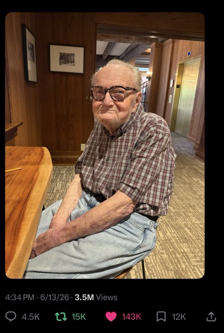 The image features an elderly man sitting at a wooden dining table, wearing glasses and a plaid shirt. The man appears to be smiling for the camera. In front of him, there are two chairs - one where he sits and another that is empty. A computer mouse can also be seen on the table next to the man's chair.