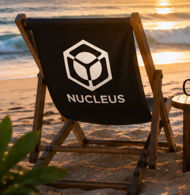A black beach chair with a wooden frame featuring the 'NUCLEUS' logo sits on a sandy beach at sunset, with ocean waves visible in the background.