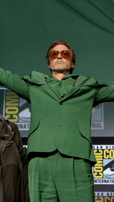 Robert Downey Jr. in a green suit with orange sunglasses poses on stage at San Diego Comic-Con, a place where major announcements like future Avengers movies are often made.