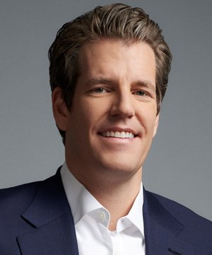 A professional headshot of a smiling man with brown hair wearing a dark suit jacket and white dress shirt against a gray background.