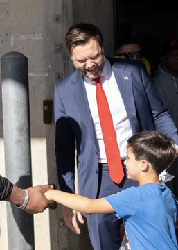 The image shows a smiling JD Vance in a suit and red tie standing beside his young son, Vivek, as the boy shakes hands with someone off-camera. It illustrates the post’s report about Vance’s son meeting Cincinnati Bengals receiver Ja’Marr Chase and being excited to be told he had nice gloves.