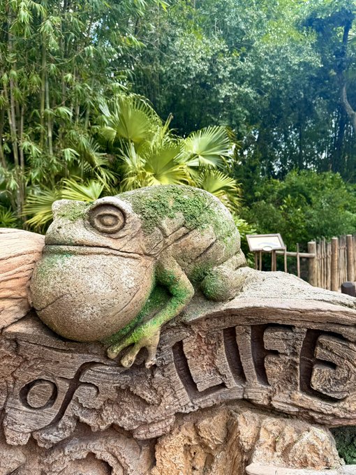 A moss-covered stone frog sculpture rests on a carved sign reading 'of LIFE' with tropical palm trees and bamboo in the background.
