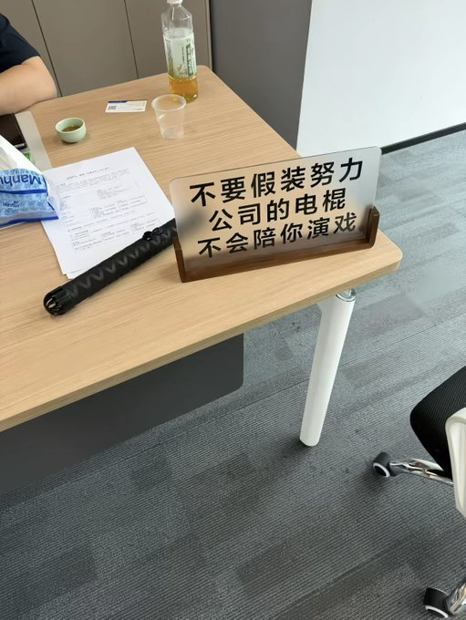 A desk sign with Chinese text reading '不要假装努力 公司的电棍 不会陪你演戏' (Don't pretend to work hard, the company's electric baton won't act with you), placed next to some papers and a bottle.