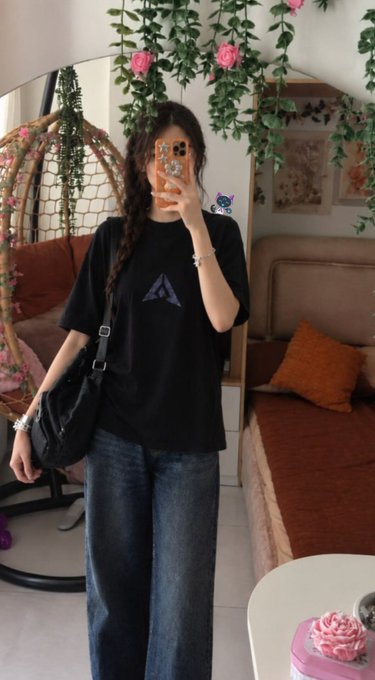 A young woman with a long braid takes a mirror selfie wearing a black t-shirt with a triangular logo and blue jeans, holding an orange phone with charms; the background features a cozy room with a hanging chair, pink flowers, and an orange bed.