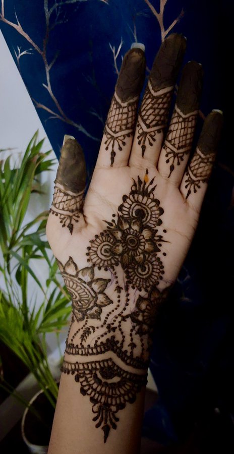 A close-up of a person's open palm displays an intricate brown henna (mehndi) design covering the hand and wrist, with dark-stained fingertips.