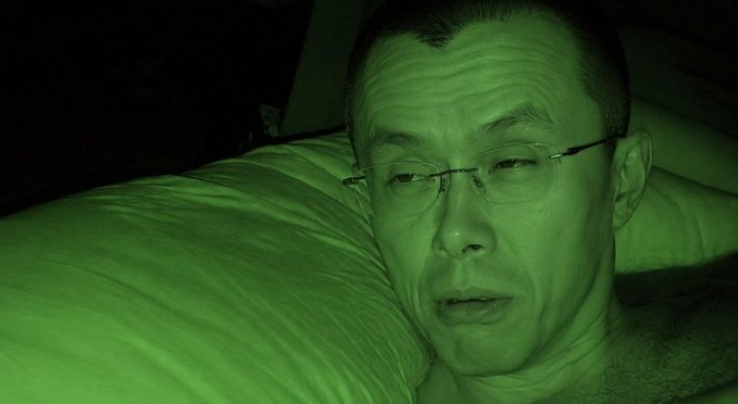 A close-up selfie of a man wearing rimless glasses lying on a pillow in a dark room with a green night-vision tint.