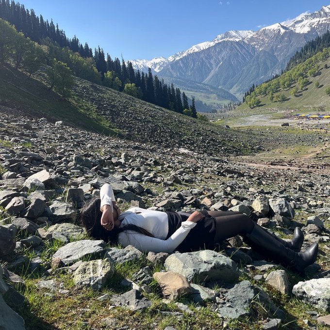 A person in a white shirt and dark pants lies on a rocky mountain slope with snow-capped peaks and green valleys visible in the background.