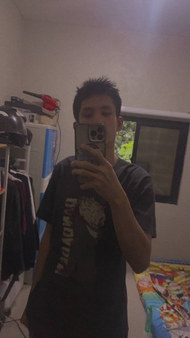 A mirror selfie of a young man taking a photo with his smartphone in a bedroom, wearing a dark t-shirt with a graphic design, with a bed featuring colorful cartoon bedding visible to the side.