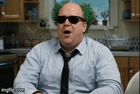 The image features a man with green hair wearing sunglasses, sitting at a kitchen counter or table. He appears to be looking off into space while wearing a tie. The background of the image shows various kitchen items such as cabinets, a sink, and a stove. There is also an "IMDb" text in the bottom right corner of the image.
