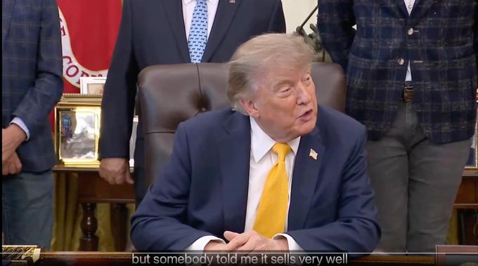 Donald Trump, wearing a dark blue suit and yellow tie, sits at a desk in what appears to be the Oval Office, speaking with the subtitle 'but somebody told me it sells very well' displayed at the bottom.