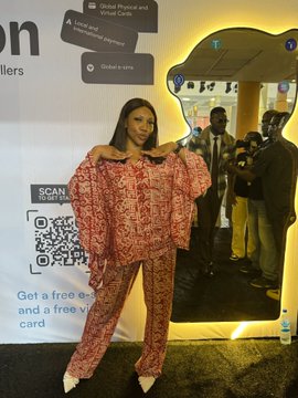 A woman poses in a red patterned off-shoulder outfit and matching pants at what appears to be a crypto or tech event, with a promotional backdrop for "SCAN ME" featuring a QR code and signage for "UPC" (likely Universal Payment Company) visible behind her. The setting aligns with the Solana Summit day 2, suggesting this image captures a moment from the cryptocurrency/blockchain conference.