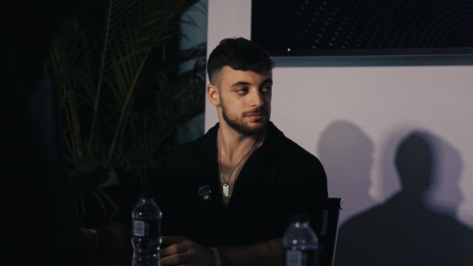 A young man with a beard sits at a table in a dimly lit setting, glancing thoughtfully to the side with water bottles visible in the foreground and plant foliage behind him, casting a relaxed, contemplative mood fitting for a "GM" (good morning) post.