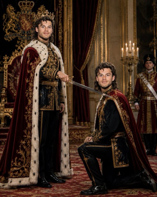 Two men in royal medieval attire; one standing wearing a crown and ermine cape holding a sword at the neck of the other kneeling man, both wearing matching red and gold outfits.