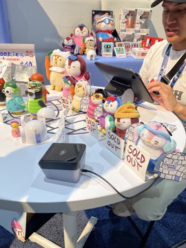 A vendor at a booth selling various small figurines and art toys, with handwritten 'SOLD OUT' signs placed among the merchandise, as a customer pays at a card reader.