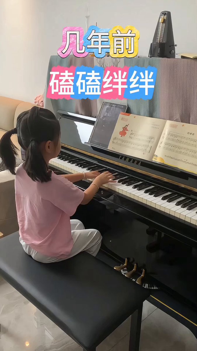 The image features a young girl sitting at a black piano, playing music with her hands on the keys. The piano has a bench next to it where she sits. A sign can be seen behind her that says "几年前还是磕磕绊绊", which translates to "Four years ago". Another sign in Chinese reads "四年后已经游刃有余", meaning "Four years have passed since the last time we saw you." The girl is sitting on a bench, and there are two books visible in the image.