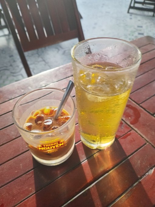 Two beverages on a wooden table: a small cup of Vietnamese iced coffee (cà phê sữa đá) with condensed milk and a tall glass of iced tea with lemon, at a street-side café.