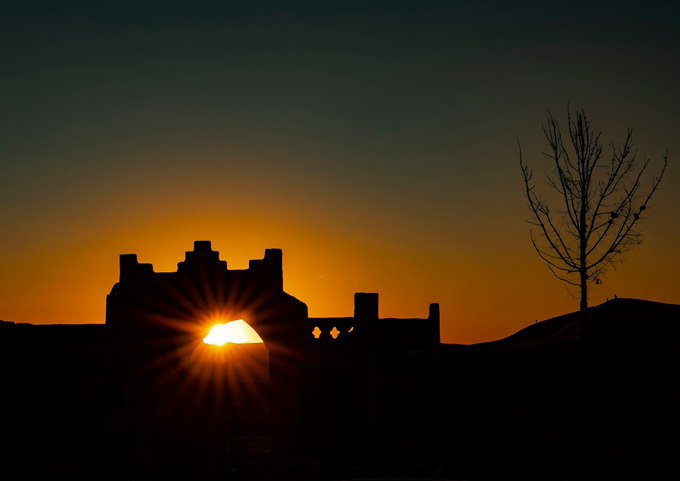 A silhouette photograph of a castle or fortress at sunset, with the bright sun creating a starburst effect through the structure, accompanied by a bare tree on the right.