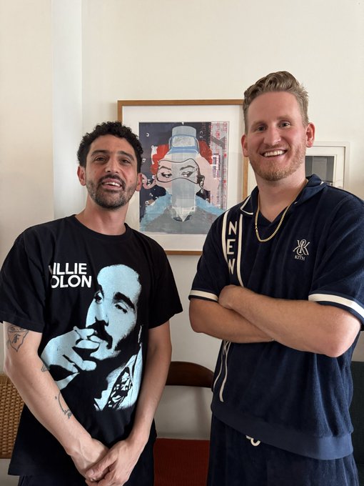 Two men posing indoors in front of a framed artwork; the left wears a black 'WILLIE COLON' t-shirt, while the right wears a navy polo shirt with 'NEV' lettering.