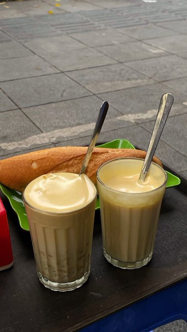 Two glasses filled with iced coffee or espresso topped with foam sit on a black table alongside bread, with spoons in the glasses.