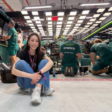 A young woman poses in an Aston Martin F1 garage during the Singapore Grand Prix, with mechanics in green "aramco" branded uniforms working on a car behind her, capturing a behind-the-scenes moment at the race that ties into the post's gratitude toward Coinbase for the experience.
