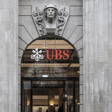 The entrance of a UBS bank building, featuring a decorative sculpted face above the arched doorway and the prominent red "UBS" logo with its three-keys symbol displayed on the glass. The image illustrates the financial institution referenced in the news about UBS significantly increasing its spot Bitcoin ETF investments to $90 million.