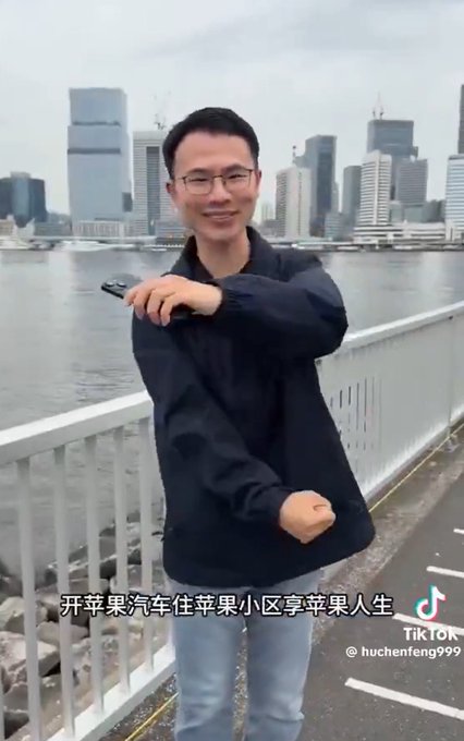 The image features a man standing next to a white railing, holding a cell phone up to his face with both hands. The man appears to be smiling for the camera while posing near the water. There are also some Chinese characters visible in the image that may provide additional context or information about the location or event taking place.