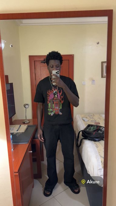A young man takes a mirror selfie in what appears to be a hotel room in Akure (as indicated by the location tag), wearing a black graphic t-shirt, black pants, and slide sandals while holding up his phone to capture the reflection. The image captures a casual "away from home" moment, tying into the post's playful reference to the lodge feeling like home—showing the cozy, simple room with a bed, desk, and wooden wardrobe that resembles lodging accommodations.